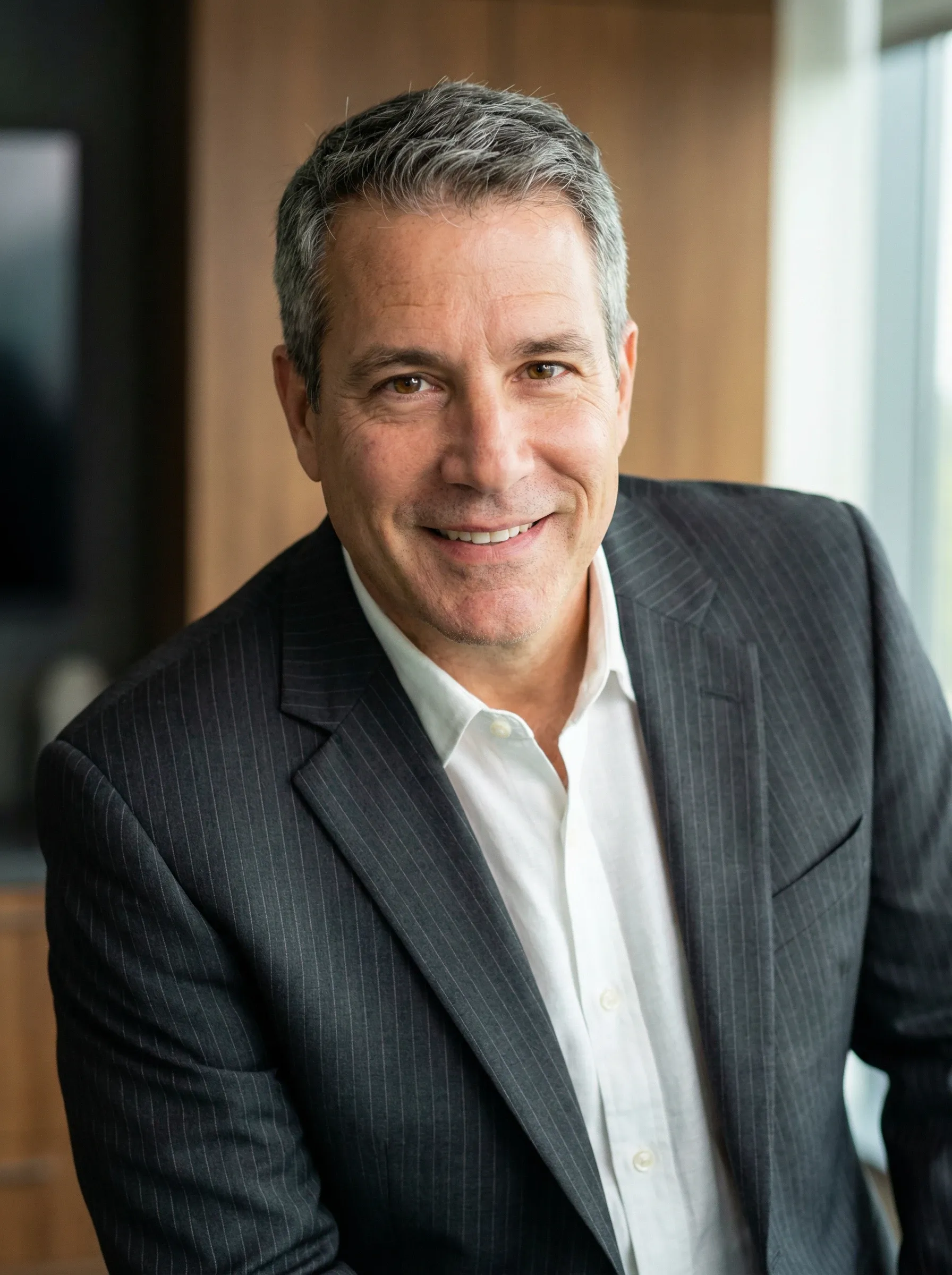 Professional headshot of a man in a charcoal pinstripe suit and white shirt in a modern office